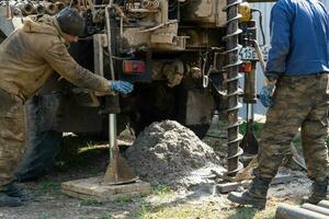 Team of workers with drilling rig on car are drilling artesian well for water in ground. Insertion of metal casing pipe into ground, installation of individual drinking supply, June 28, 2022, Russia, photo