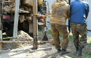 Team of workers with drilling rig on car are drilling artesian well for water in ground. Insertion of metal casing pipe into ground, installation of individual drinking supply, June 28, 2022, Russia, photo