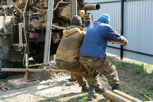 Team of workers with drilling rig on car are drilling artesian well for water in ground. Insertion of metal casing pipe into ground, installation of individual drinking supply, June 28, 2022, Russia, photo