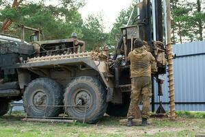 Team of workers with drilling rig on car are drilling artesian well for water in ground. Insertion of metal casing pipe into ground, installation of individual drinking supply, June 28, 2022, Russia, photo