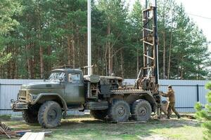 Team of workers with drilling rig on car are drilling artesian well for water in ground. Insertion of metal casing pipe into ground, installation of individual drinking supply, June 28, 2022, Russia, photo