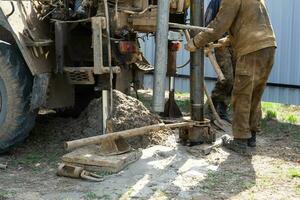 Team of workers with drilling rig on car are drilling artesian well for water in ground. Insertion of metal casing pipe into ground, installation of individual drinking supply, June 28, 2022, Russia, photo