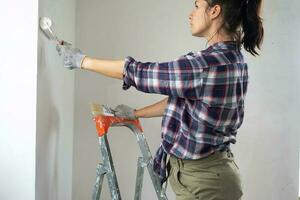 Woman in paint roller and white paint paints the wall of house on stepladder. Construction work and cosmetic repairs in house, wall painting, tinting, finishing work with your own hands photo