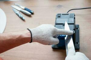 afilando un cuchillo en un afilador eléctrico en casa. la mano del hombre conduce la hoja del cuchillo entre los afiladores azules, el polvo vuela sobre la máquina. foto