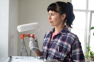 puzzled woman in paint roller and white paint for walls in hands close-up portrait. Construction work and cosmetic repairs in house, wall painting, tinting, finishing work with your own hands photo