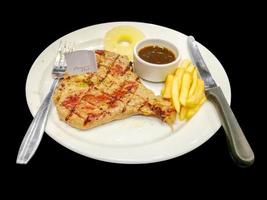 T-bone pork steak with french fries and pine apple, pepper sauce on white ceramic plate with steak knifes and fork isolate on black background and make with paths. photo