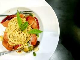 Top view sausage spaghetti in Thai style on white ceramic plate with spoon and fork on black leather table in the restaurant. photo