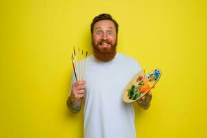 Happy man with beard and tattoo is ready to draw with brushes photo