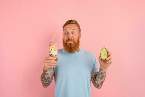 Thoughtful man with beard and tattoos is undecided if to eat an icecream or an avocado photo