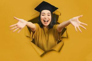 contento asiático chica, con graduación sombrero, viene fuera de un agujero en un amarillo papel fondo foto