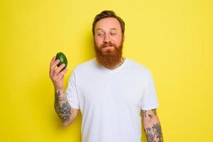 Happy man with beard and tattoos holds an avocado photo
