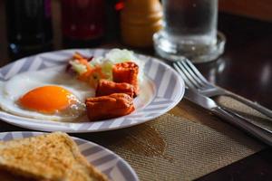 Breakfast, fried eggs, fried sausage, vegetable salad and toast on a brown wooden table with coffee. photo