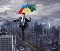 Equilibrist businessman walk on a rope with umbrella over the city. Concept of overcome the problems and positivity photo