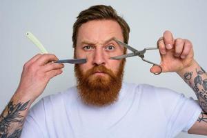 Worried man with scissors and blade is ready to cut the beard photo