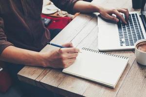 Young business man hand writing notebook and using laptop on wood table. photo