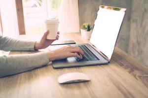 Woman holding coffee and using laptop on wooden table in office photo