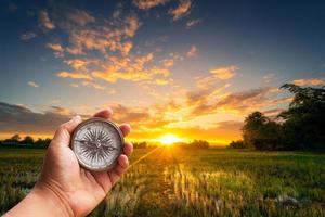 un hombre participación Brújula en mano a campo y puesta de sol para navegación guía. foto