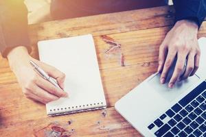 Asia business man hand using laptop and writing note pad on table in coffee shop with vintage toned filter. photo