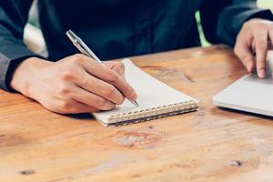 Man hand writing notebook paper and using phone on wood table in coffee shop with vintage toned filter. photo