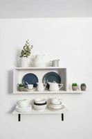 many ceramic and porcelain dishes stand on white open kitchen shelves with indoor flowers in pots. stylish interior view of part of the kitchen. photo