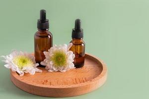 two cosmetic bottles with a dark glass pipette with a natural remedy, oil, serum for personal care on a wooden tray with flowers. green background. photo