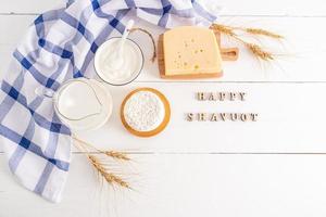 fresh dairy products, apples, a bouquet of ears of corn on a white wooden background with wooden letterstext of happy shavuot. top view. photo