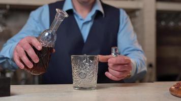 A Bartender in a blue shirt and black vest stands behind the Bar, takes a Decanter with Whiskey Pouring into a glass. Close-up video