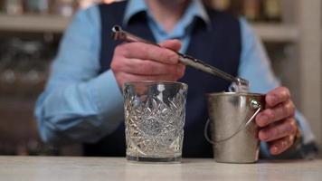 Bartender with steel tongs Puts Ice in Whisky Glass. Elegant bartender in blue shirt and black vest stands behind Bar counter and makes Cocktail. Close up video