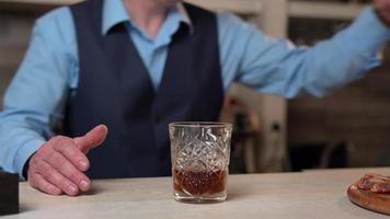 A Close-up of the hands of the Bartender, that Removes the Whisky Bottle from the Bar counter. A Bartender in a blue shirt and black vest stands behind the Bar counter Preparing a Cocktail video