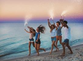 contento sonriente amigos corriendo a el playa con espumoso velas foto