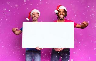 Surprised Amazed friends with Christmas hat hold a white banner for your message. Purple background photo