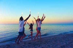 contento sonriente amigos corriendo a el playa con espumoso velas foto