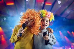 dos niño canta un canción con micrófono y gracioso peluca foto