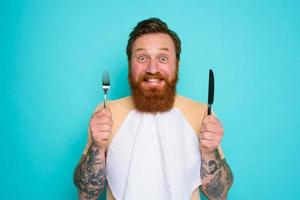 Happy man with tattoos is ready to eat with cutlery in hand photo