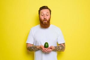 Amazed with beard and tattoos holds an avocado photo
