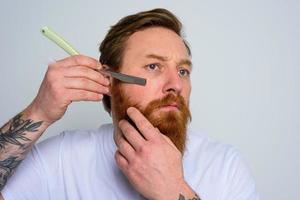 Serious man with blade is focused on cutting his beard photo