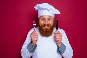 happy chef with beard and red apron holds cutlery in hand photo