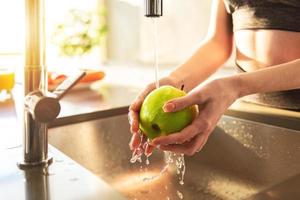 joven mujer lava, con corriendo agua, un manzana en el cocina lavabo iluminado por el Dom foto