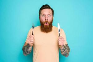 Man with tattoos is ready to eat something with cutlery in hand. cyan background photo