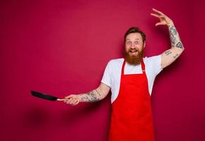 Happy chef with beard and red apron cooks with pan photo