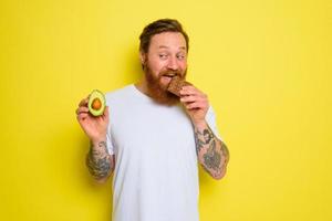 escéptico hombre con barba y tatuajes sostiene aguacate y chocolate foto