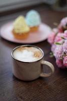 A Close-up of a beige Cappuccino Cup and Two yellow and blue Cream Capcakes on a pink plate sit on a wooden table. Near a plate of cakes lie beautiful pink and white flowers. photo