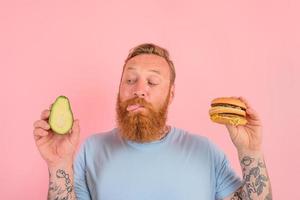 Hungry man with beard and tattoos is undecided if to eat an avocado or an hamburger photo