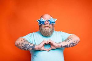 grasa hombre con barba, tatuajes y Gafas de sol hace corazón forma con manos foto