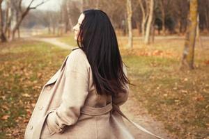 portrait of brunette hair woman in beige coat walking at the city park . photo