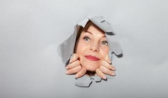 Surprised woman looking playfully in torn paper hole, has excited cheerful expression, looks through breakthrough of gray background. Wow, great news photo