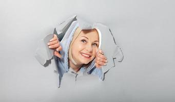 Charismatic joyful woman with blonde hairstyle, looking playfully, wears fashionable hoody, stands in torn paper hole, notices funny pleasant. Gray background. photo