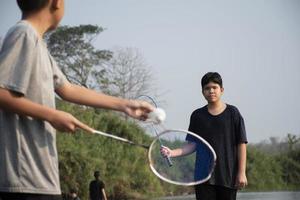 asiático Niños sostener bádminton volante y raqueta, en pie y jugando junto a el río banco en su local río durante su fin de semana día festivo, suave y selectivo atención en frente chico en blanco camisa. foto