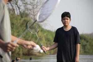 Asian boys hold badminton shuttlecock and racket, standing and playing beside the river bank in their local river during their weekend holiday, soft and selective focus on front boy in white shirt. photo