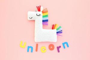 Crochet amigurumi handmade stuffed soft white unicorn toy with rainbow mane and word on pink background. Handwork hobby. Craft diy newborn pregnancy concept. Knitted doll for little baby. Flat lay photo